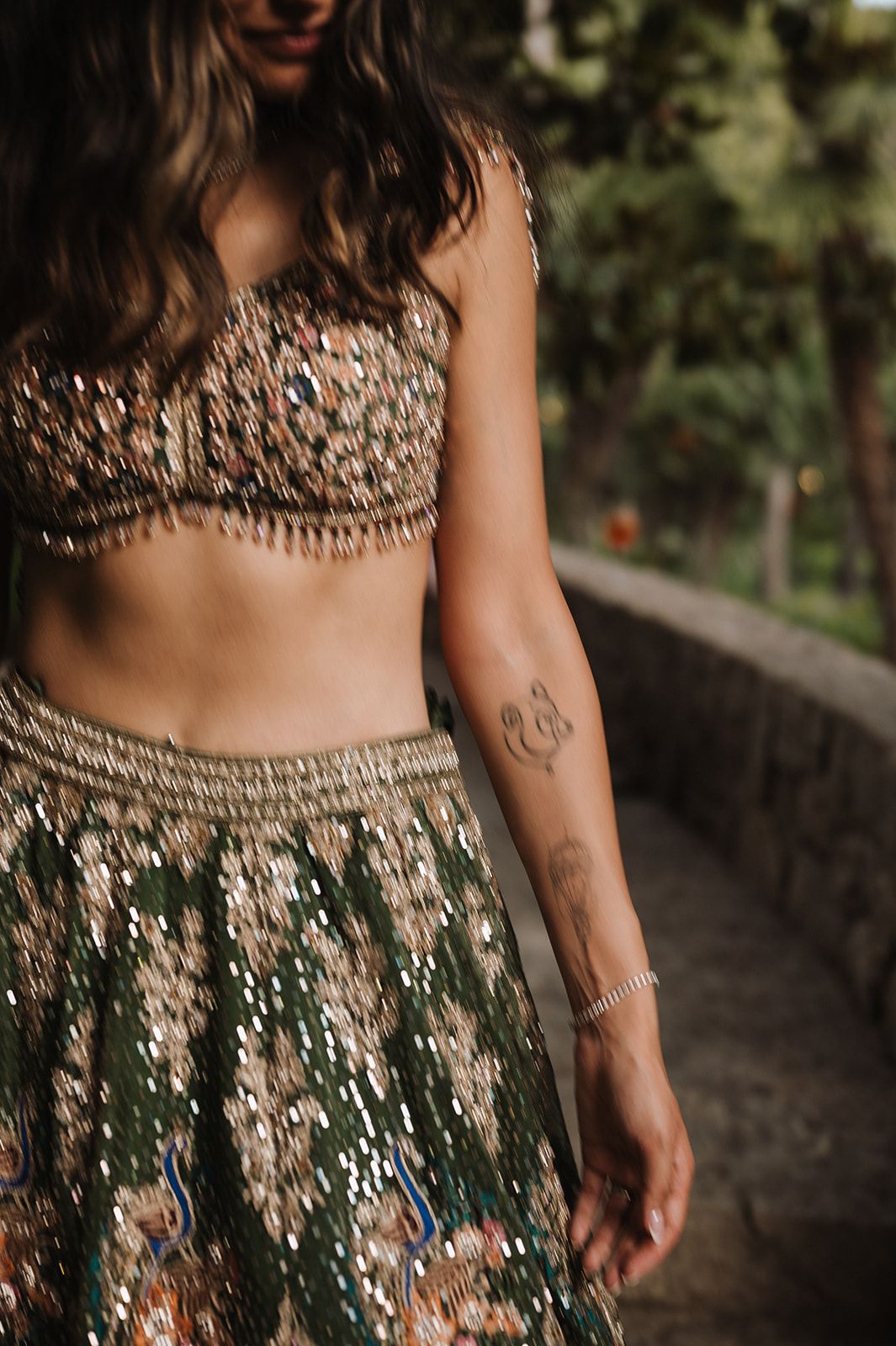 Woman in embellished crop top and skirt with tattoo at outdoor wedding event.