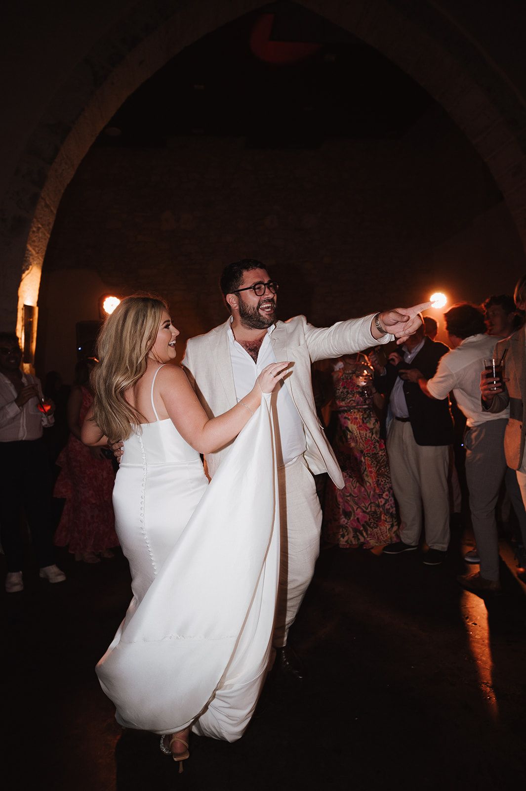 Bride and groom dancing energetically under stone arch at wedding reception with guests watching behind them.