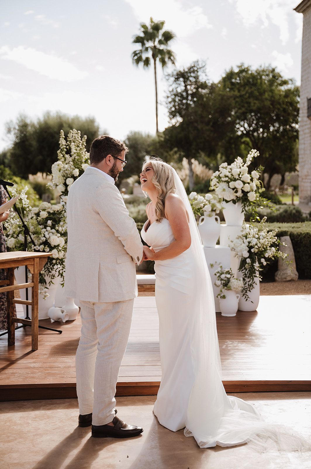 Bride and groom exchanging vows at an outdoor ceremony with white floral arrangements and a palm tree backdrop.
