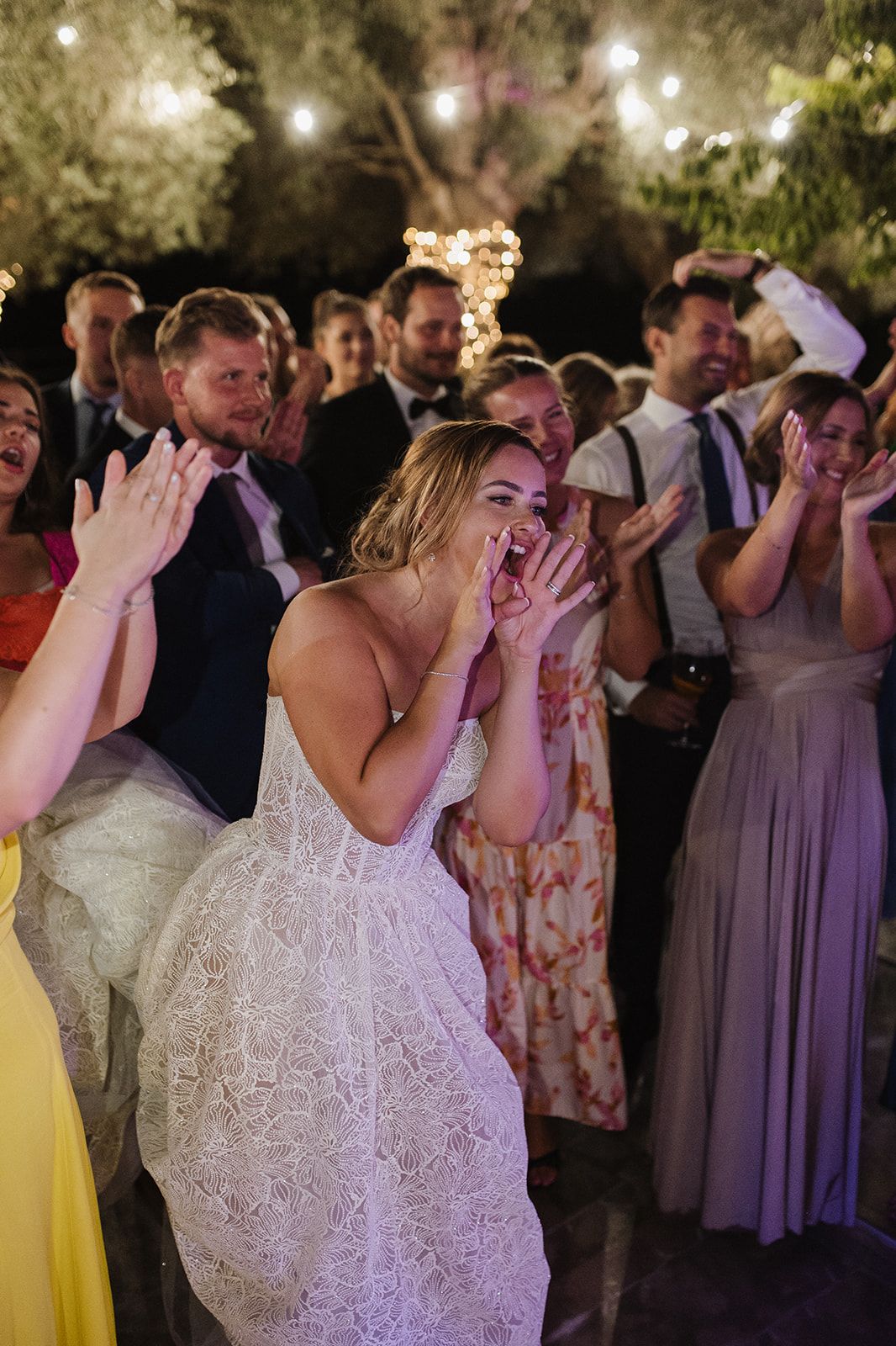Excited bride in lace gown cheering with hands on face surrounded by guests at reception.