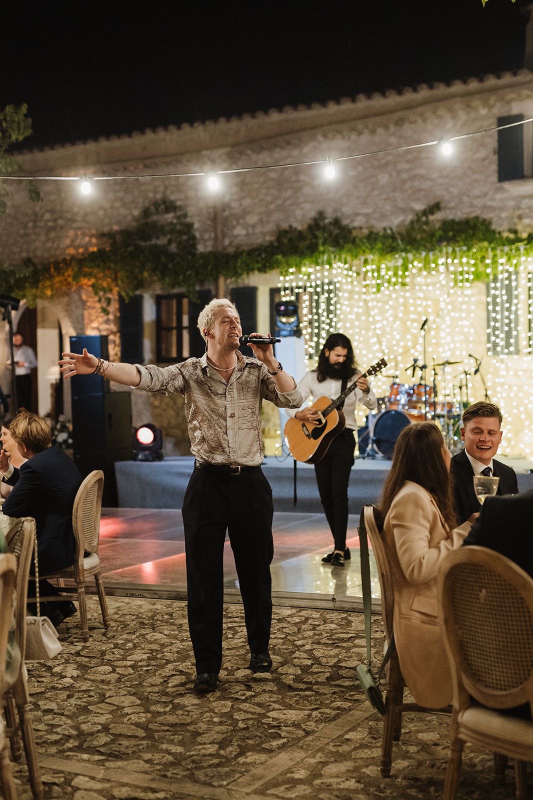 Wedding entertainer performing with open arms while live band plays under string lights at night.