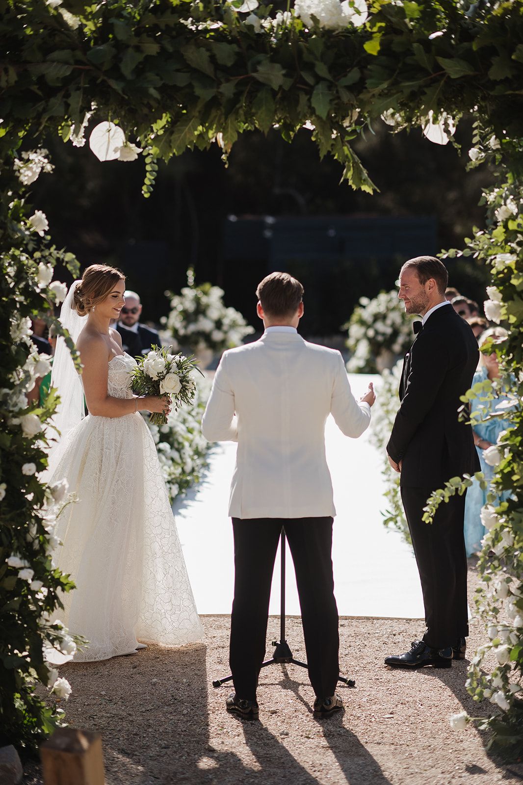 Bride and groom exchange vows under a lush green floral arch at an elegant outdoor wedding ceremony.