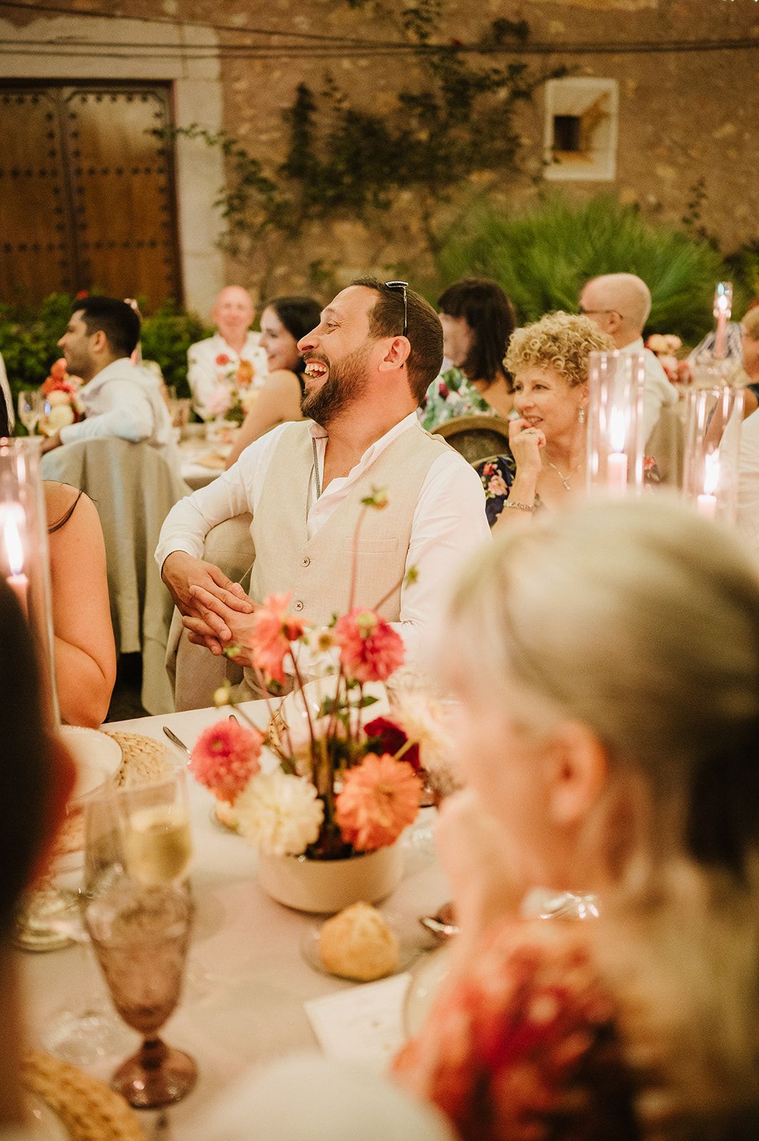 A laughing man in a linen vest enjoys a wedding reception dinner with floral centerpieces and candles.