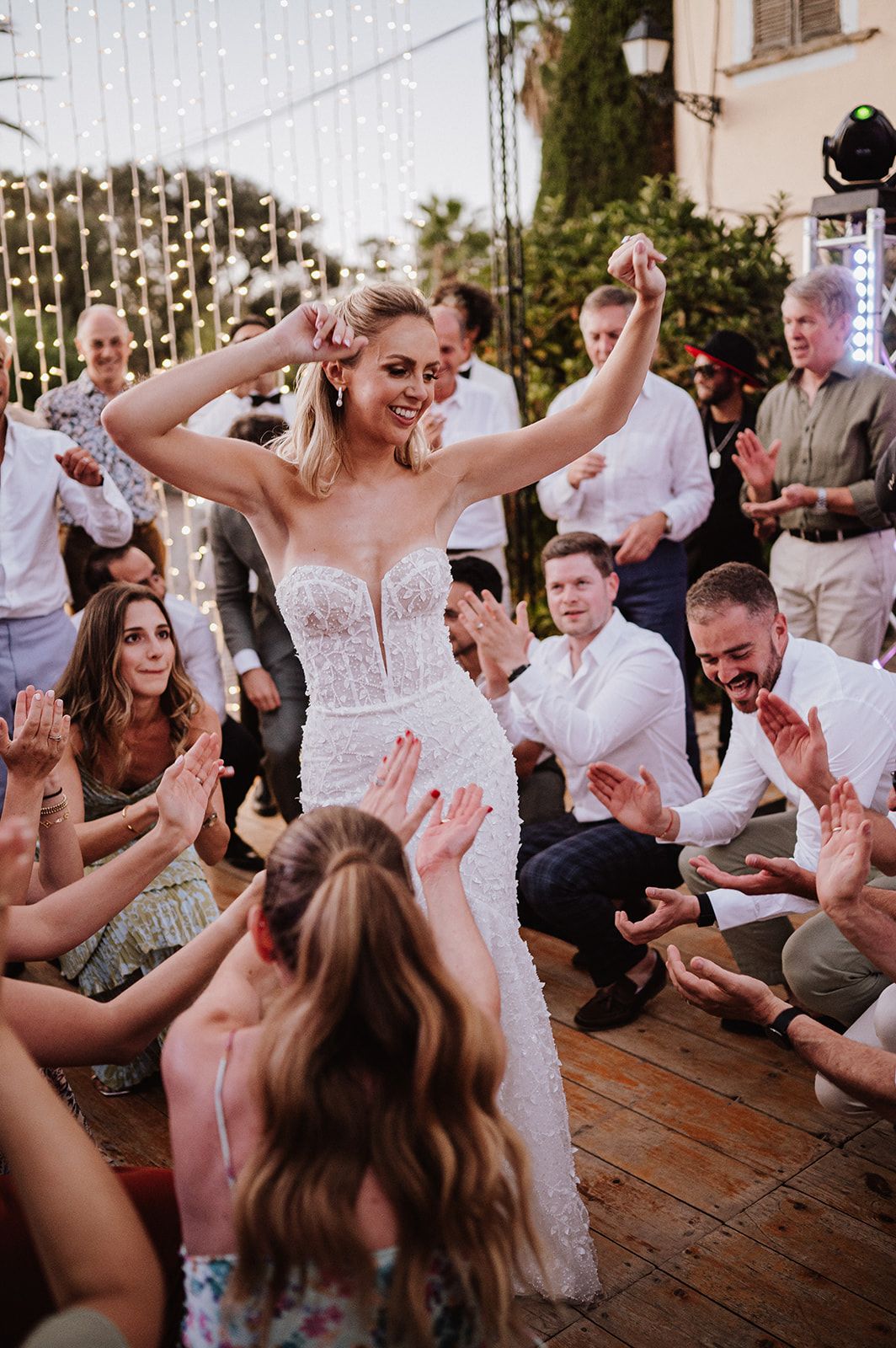 Bride in fitted lace gown dances joyfully surrounded by cheering guests at outdoor reception.