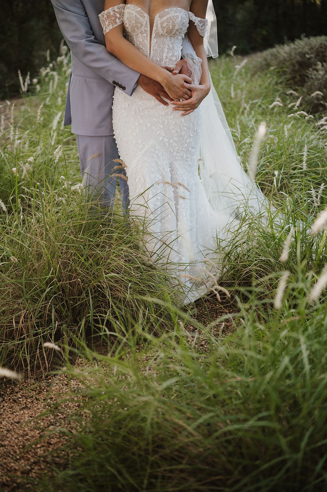 Bride in lace gown embraced by groom in gray suit among tall grass.