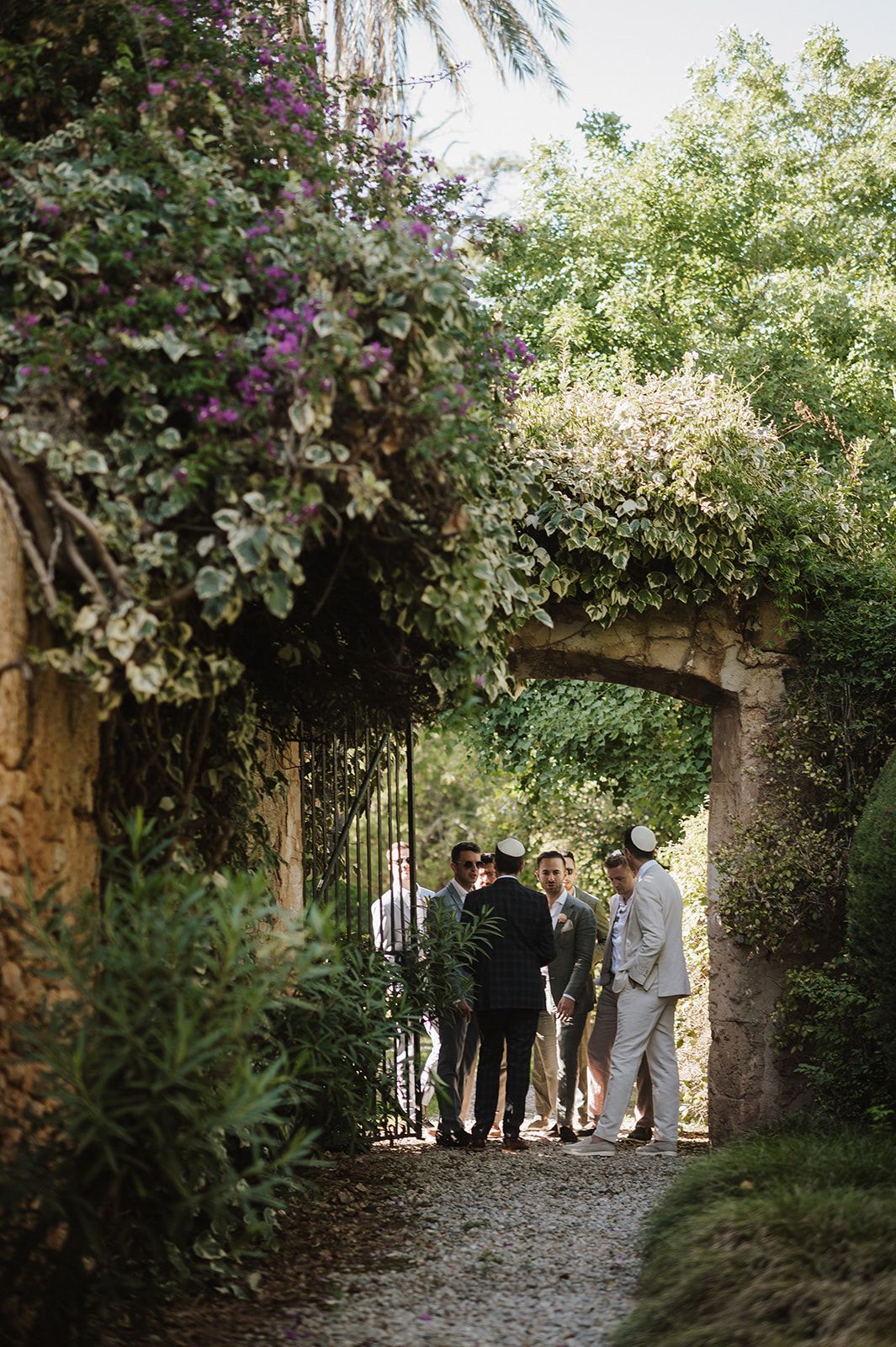 Wedding guests gather under a flower-covered stone archway surrounded by lush greenery at an outdoor venue.