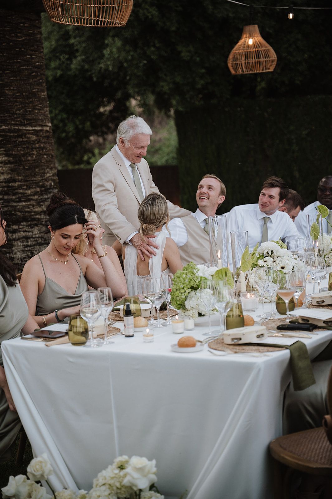 Elegant outdoor wedding reception dinner with guests laughing at a long white-clothed table under wicker pendant lights.