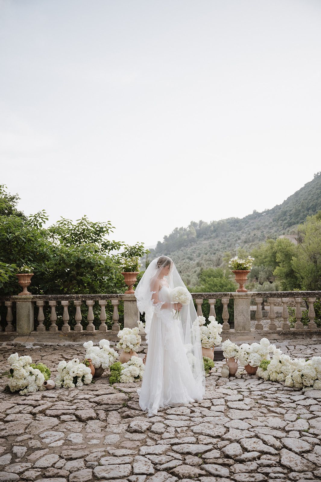 Bride in flowing white gown and veil stands among white hydrangea arrangements on a rustic stone terrace.