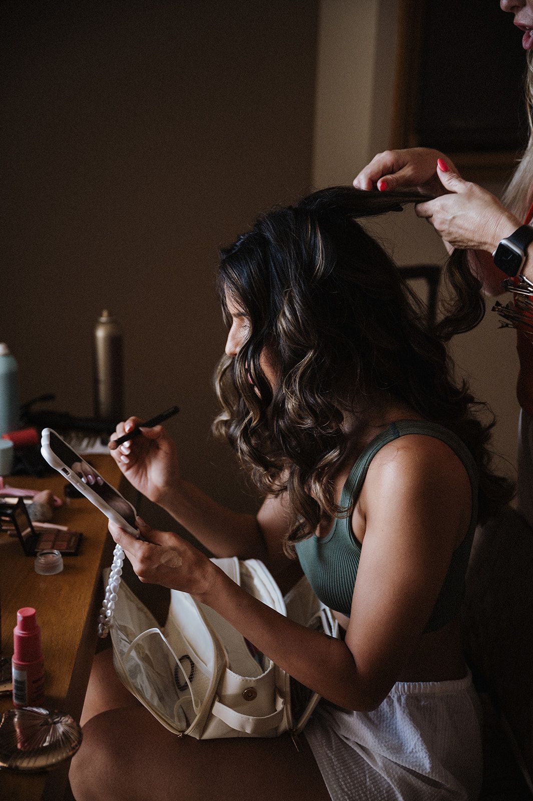 Bride applying makeup while getting hair styled with curling iron before Indian wedding ceremony.