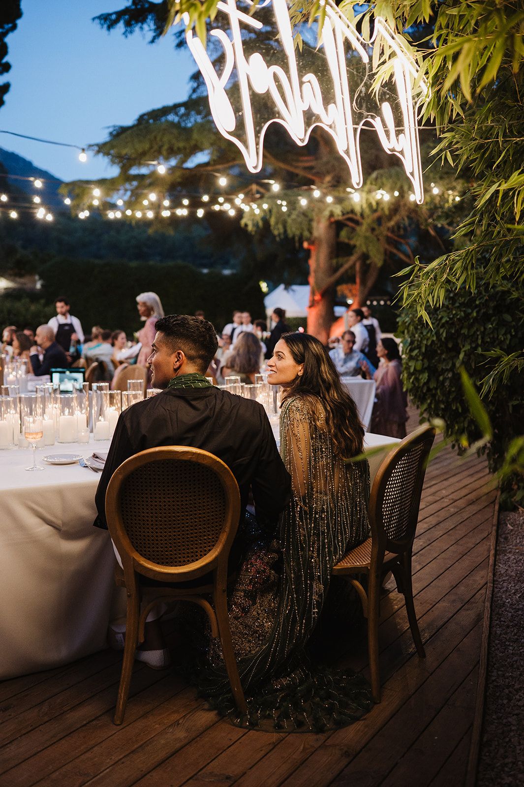 Bride and groom sit at candlelit reception table under glowing string lights and neon wedding sign.