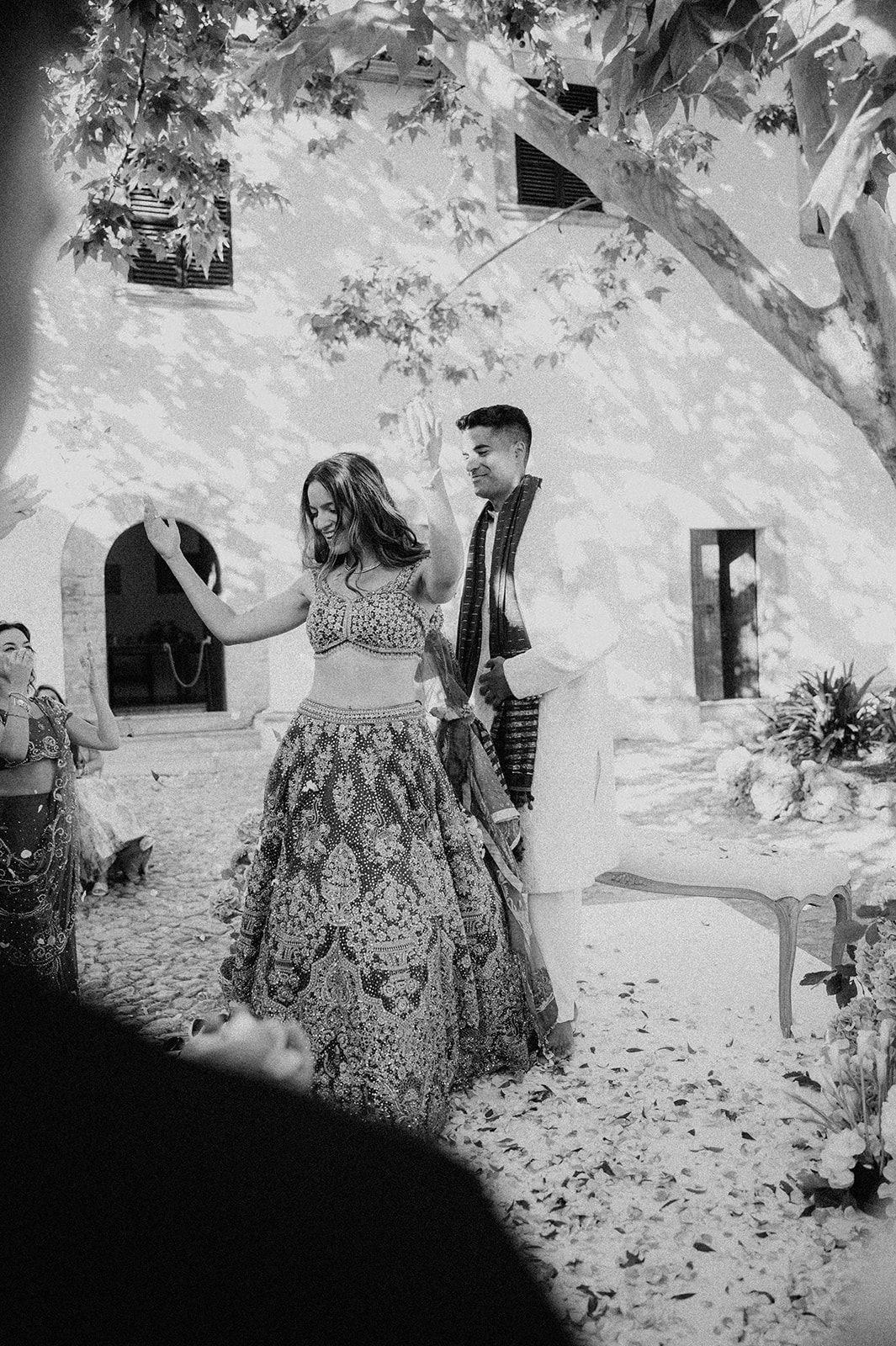 Black and white photo of bride in embroidered lehenga dancing with groom at Indian wedding ceremony.