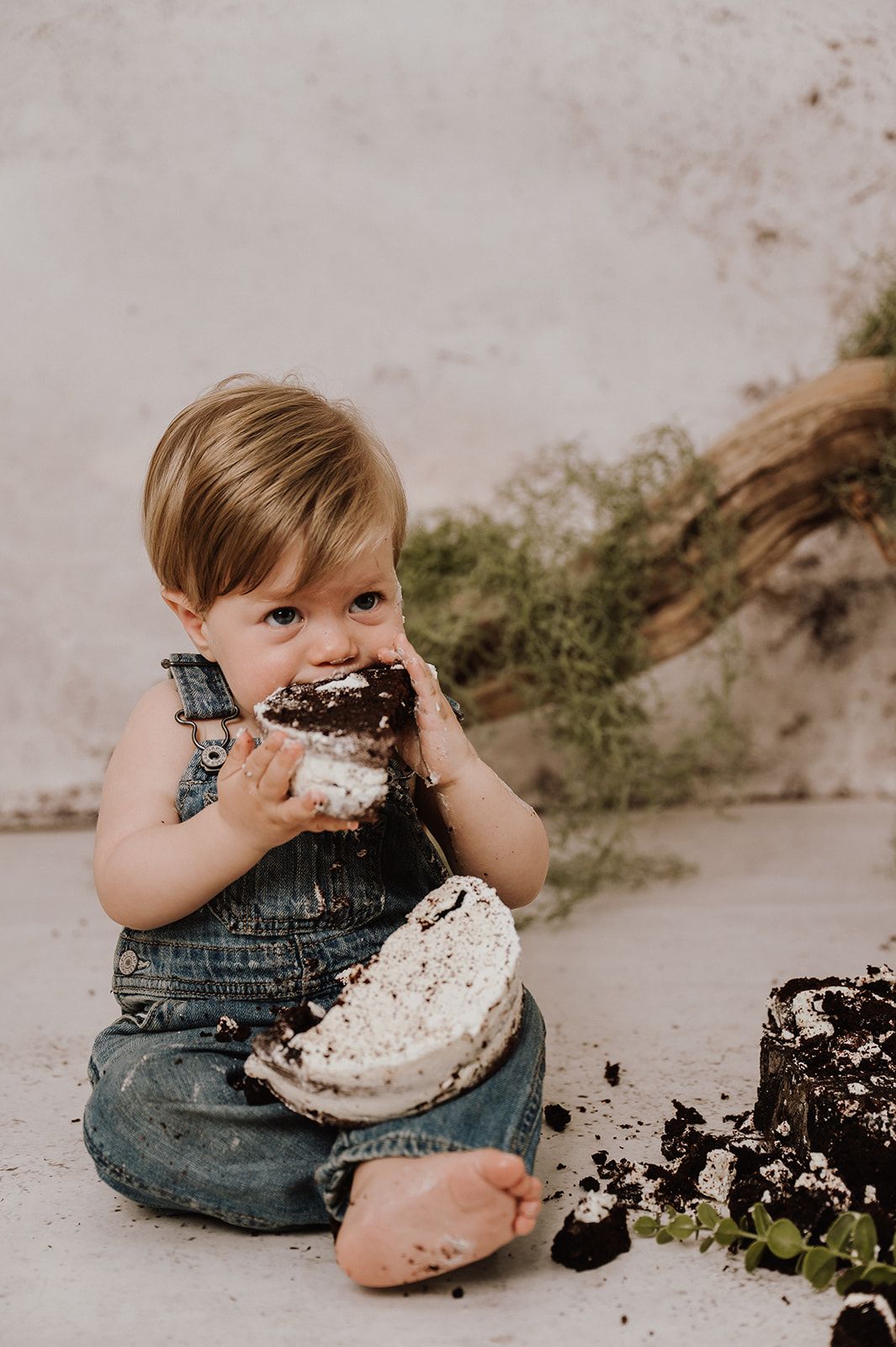 Toddler in denim overalls enjoying a cake smash, covered in chocolate frosting during first birthday photos.