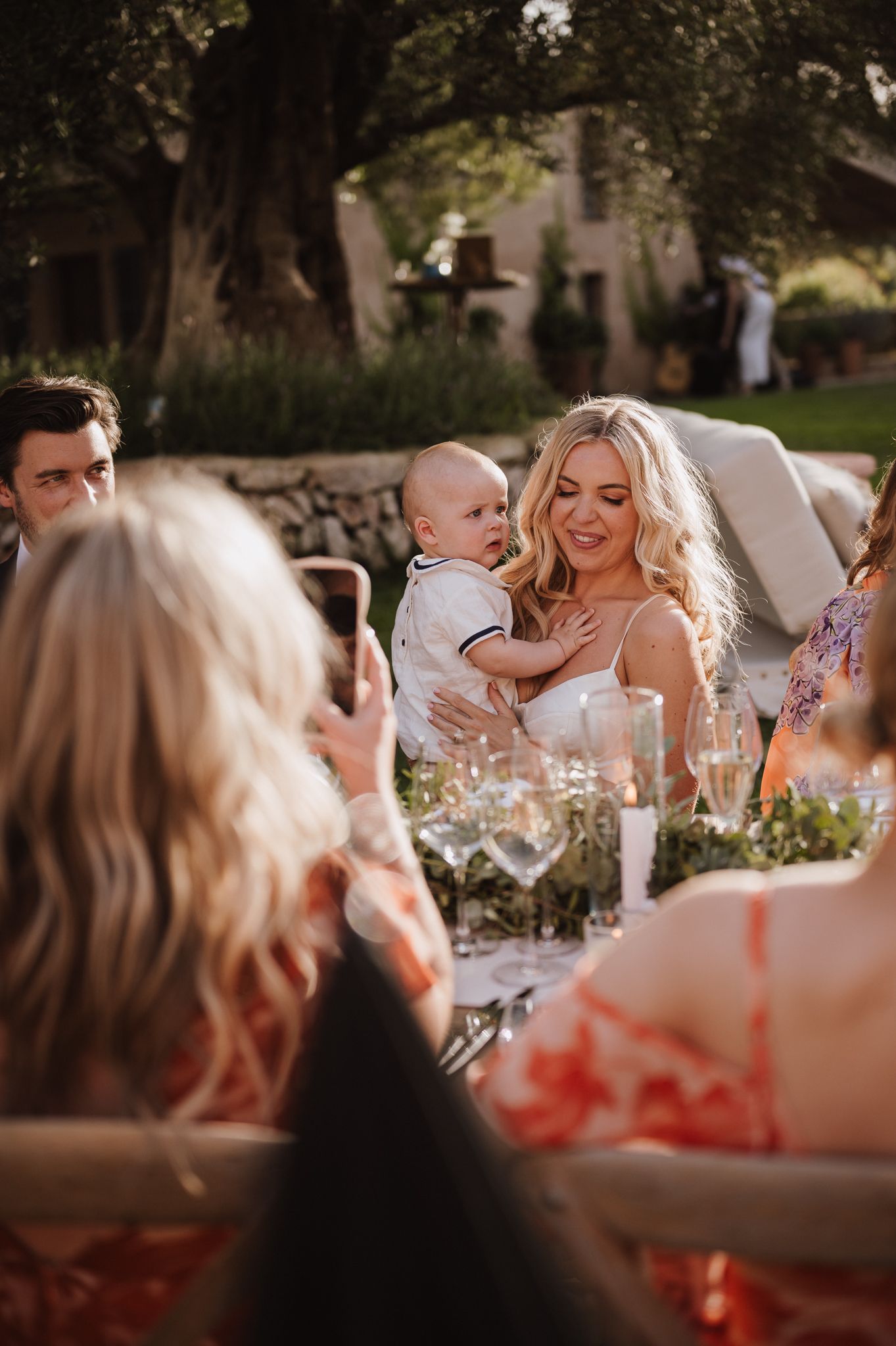 Smiling blonde bride in white dress holding an infant at a sunlit outdoor wedding reception dinner table.
