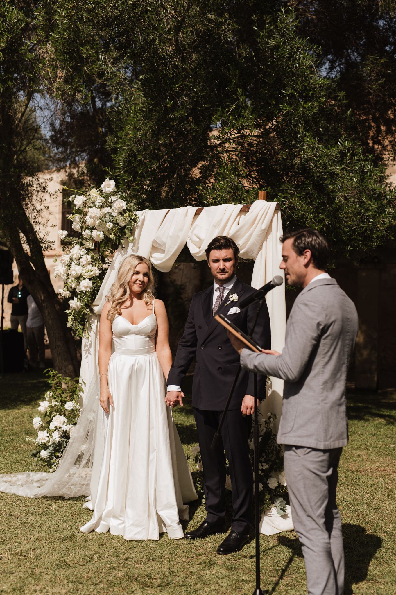Bride and groom exchange vows at outdoor altar adorned with white flowers and draped fabric with officiant.