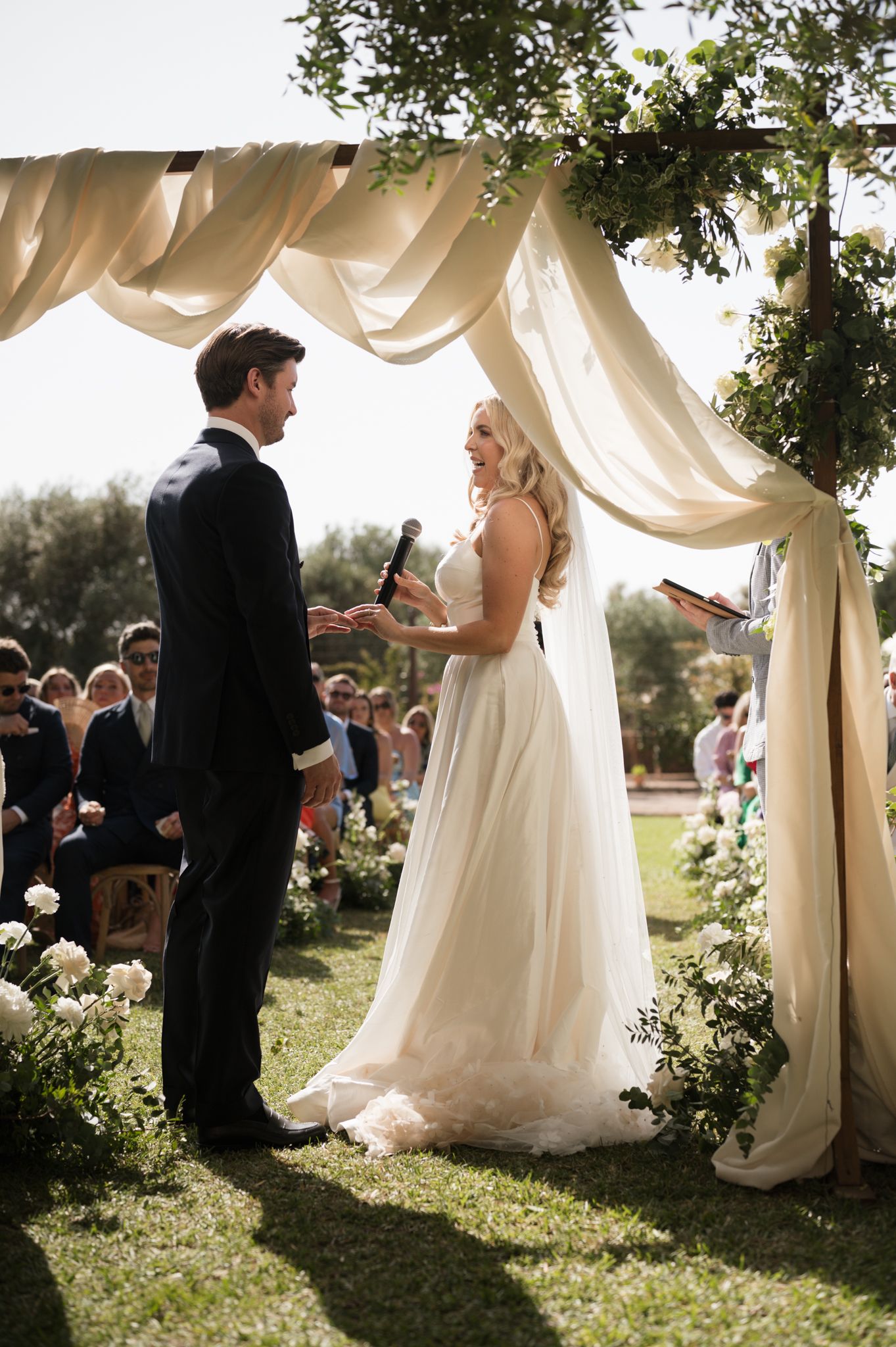 Bride reading vows to groom under elegant draped fabric wedding arch surrounded by white floral arrangements.