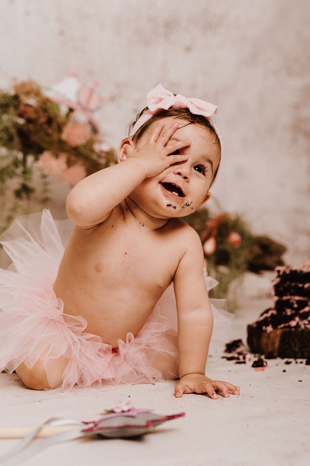 Baby girl in pink tutu smearing chocolate cake on her face during first birthday cake smash photo session.