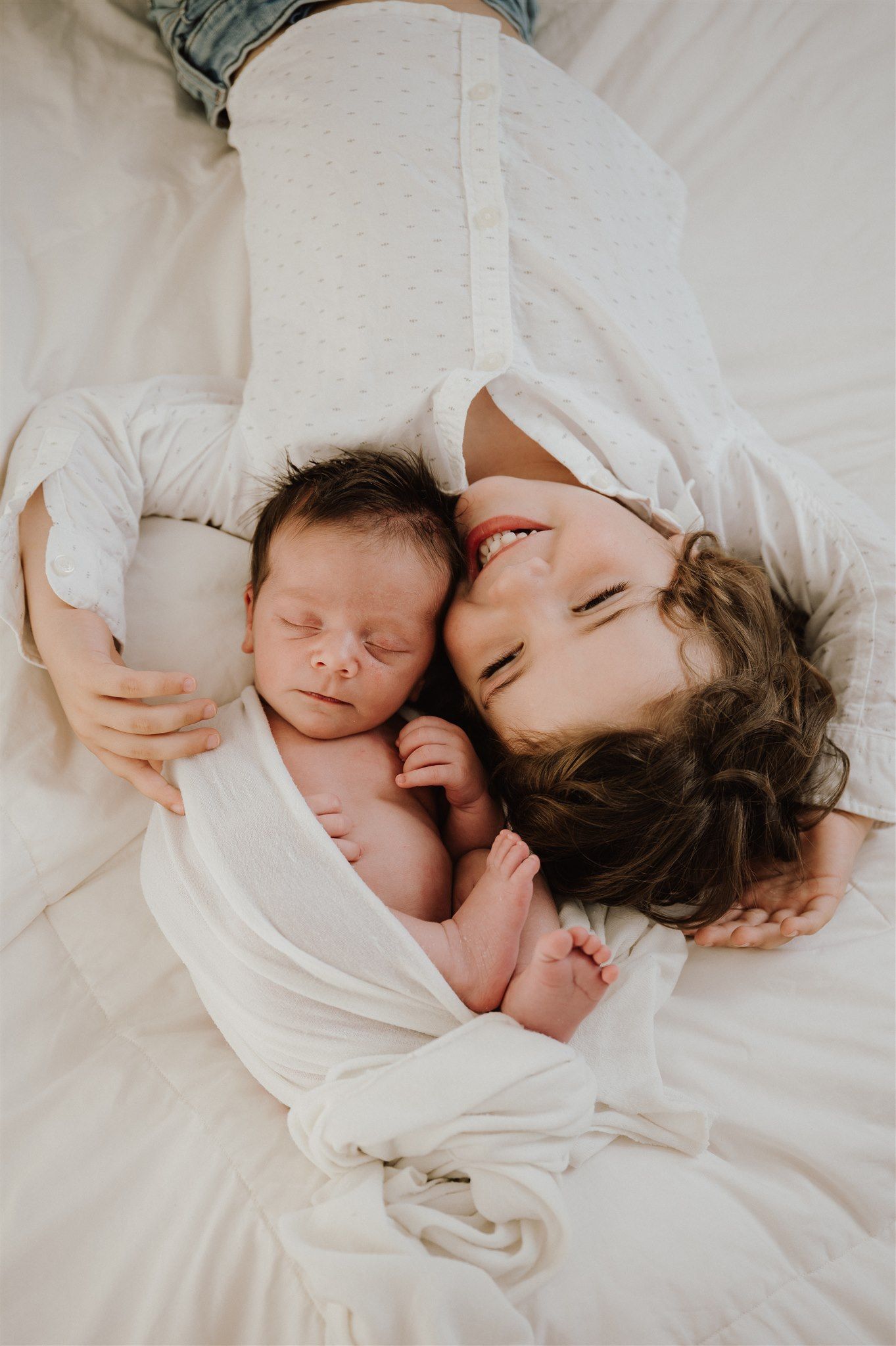 A young child smiles while lying beside a sleeping newborn baby, both wrapped in white on a soft bed.