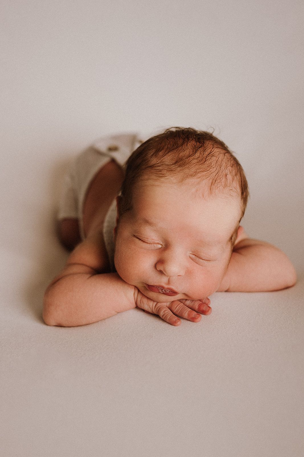 Peaceful newborn baby sleeping with hands tucked under chin, wearing a cream romper on a soft white background.