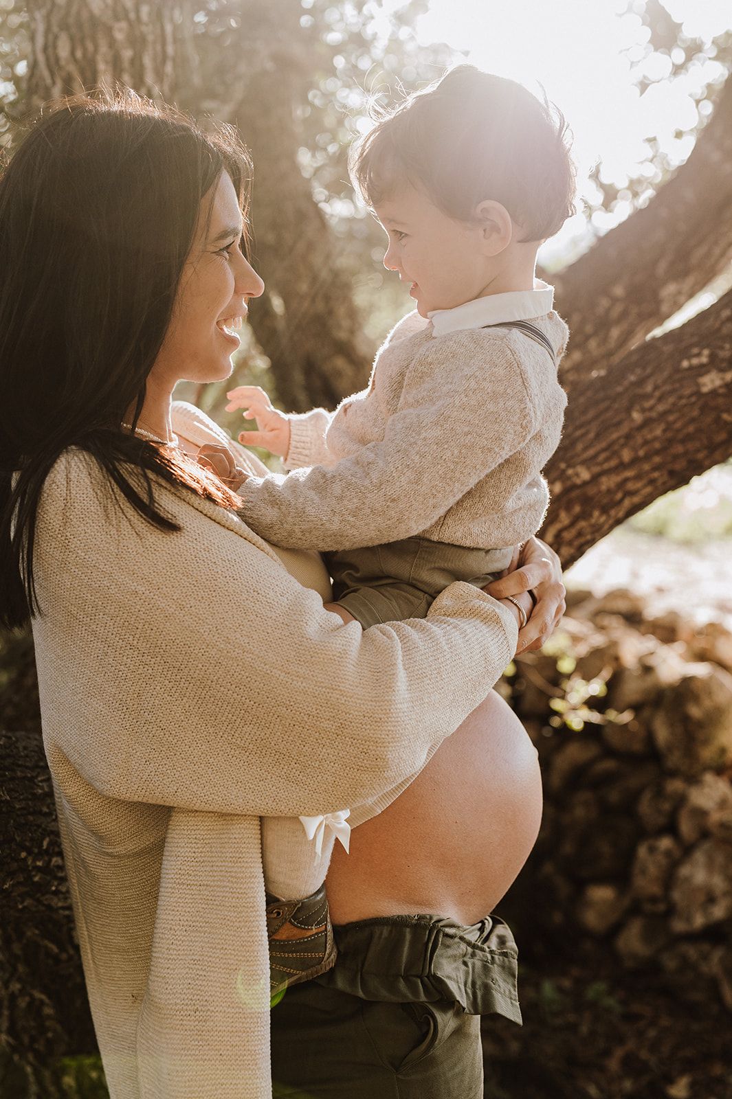 Pregnant woman holding toddler son outdoors in golden sunlight near trees, wearing beige sweater.