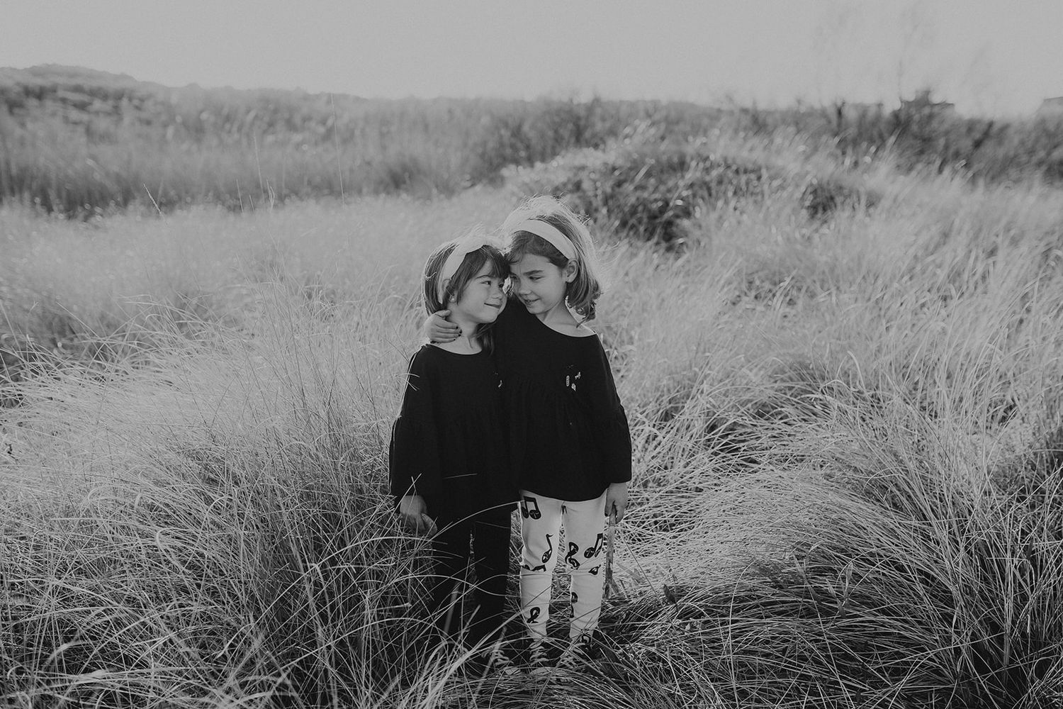 Black and white photo of two young siblings in hats standing close together in tall grass fields.