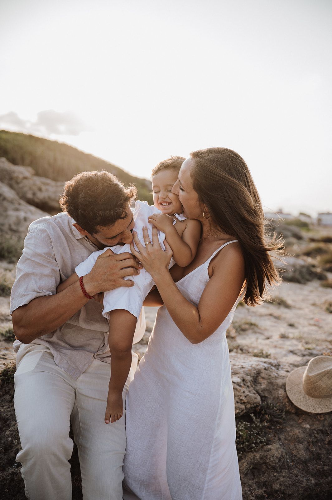 Happy family with parents kissing their laughing baby on a sunny rocky beach.
