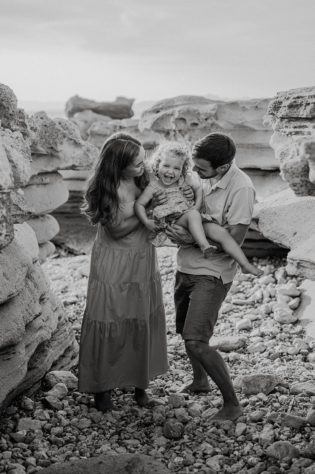 Black and white photo of parents laughing with their toddler on a rocky beach.