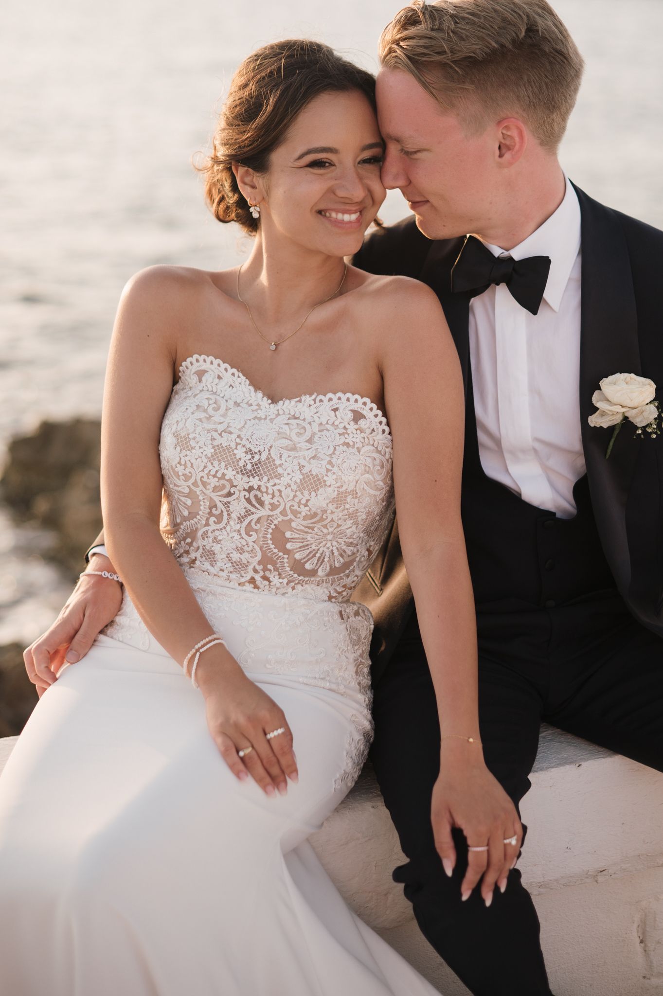 Smiling bride in strapless lace gown and groom in tuxedo pose by the sea.