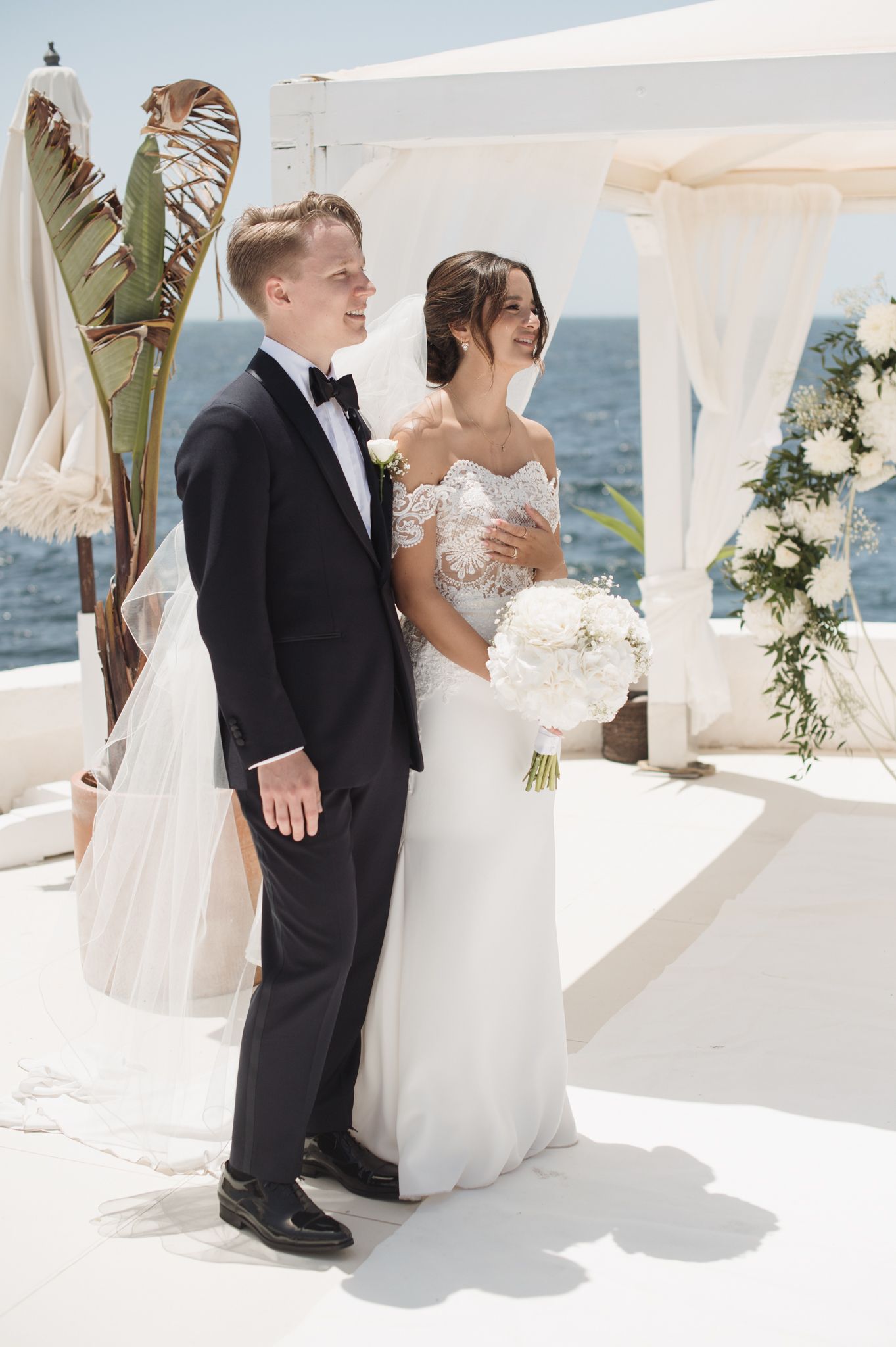 Bride holding white bouquet stands with groom at oceanfront wedding venue.