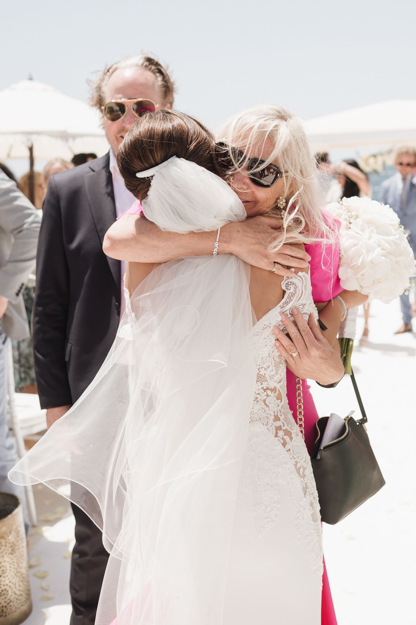 Bride in lace wedding dress receives warm hug from guest in pink at outdoor wedding ceremony.