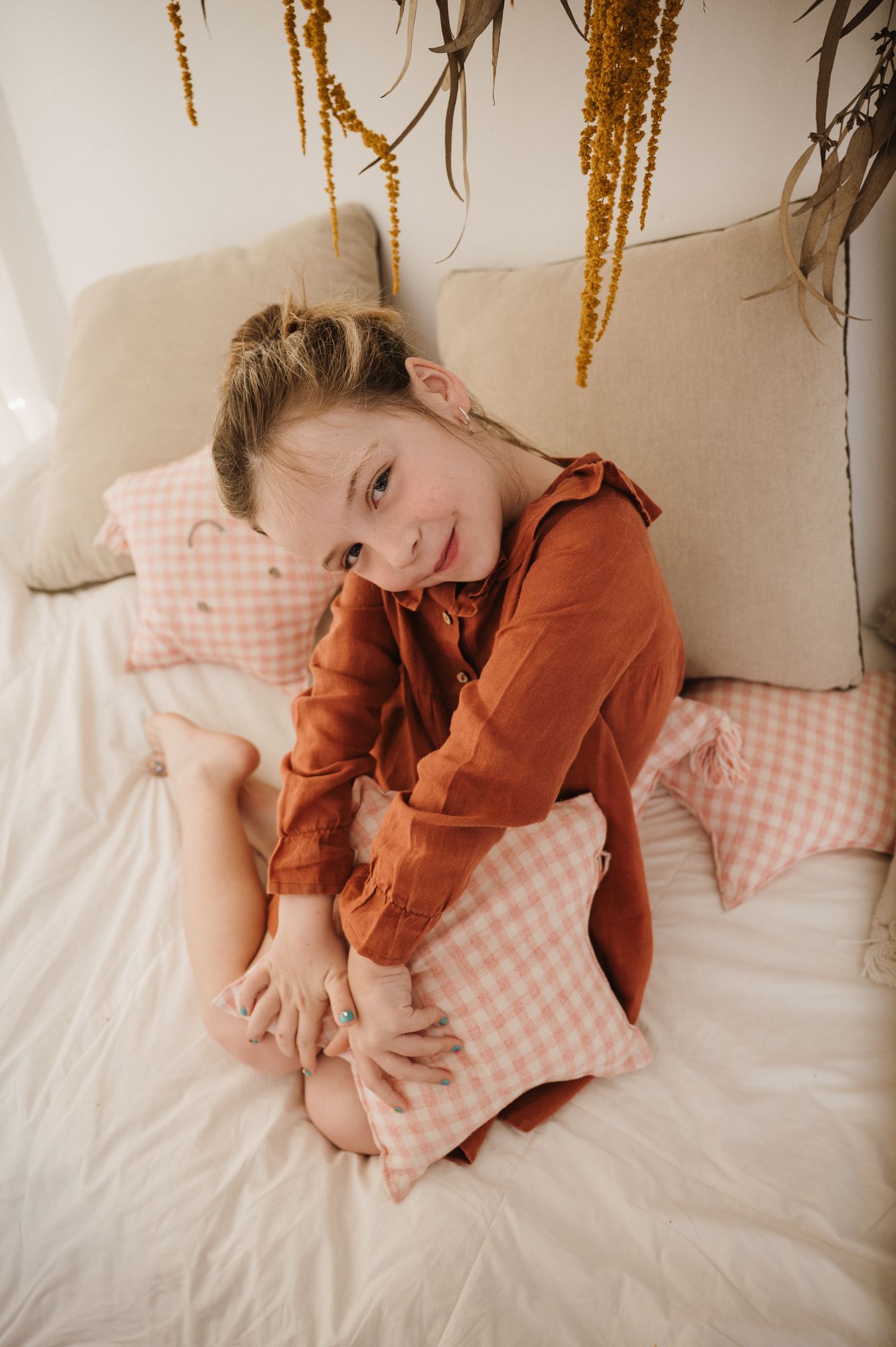 Young girl in rust dress hugging pink gingham pillow on white bed with dried flowers.