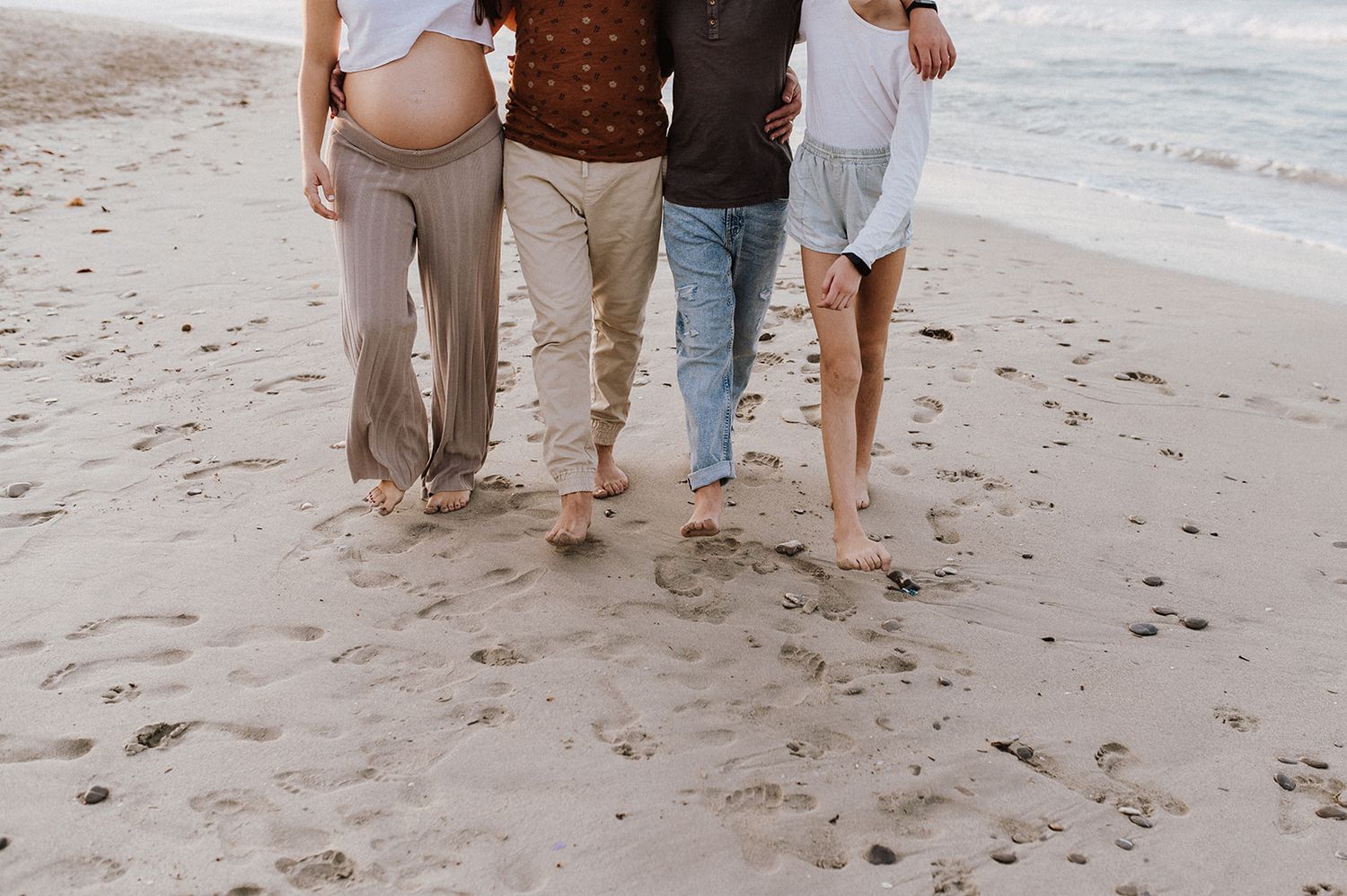 Three barefoot people walking on sandy beach, pregnant woman in neutral tones, ocean waves in background.