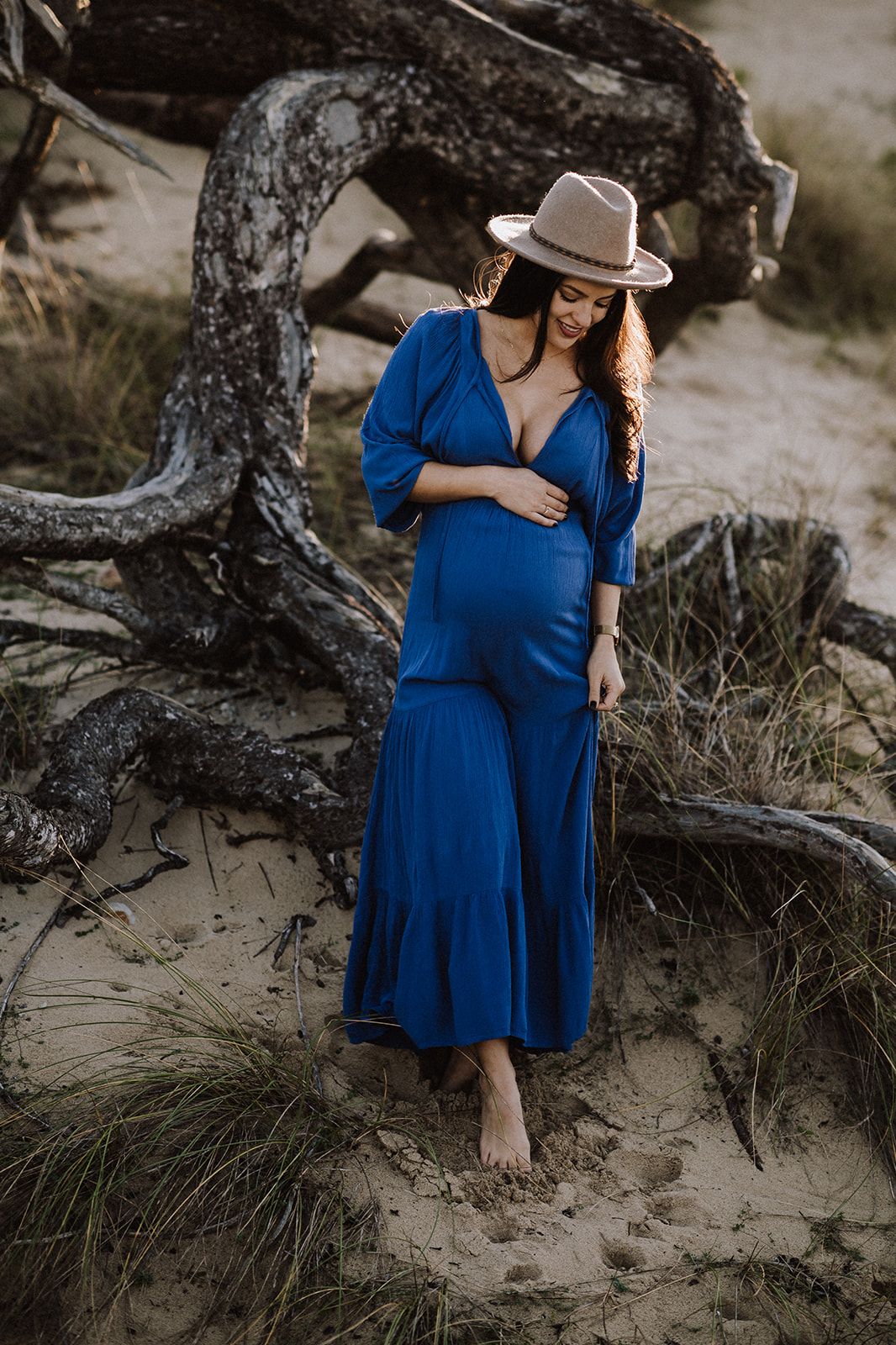 Pregnant woman in blue maxi dress and tan hat stands barefoot on sandy beach near driftwood.