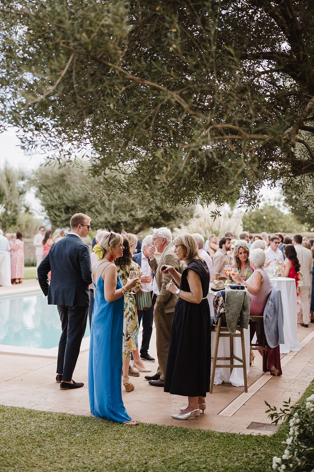 Elegant outdoor wedding cocktail hour with guests mingling beside a pool under olive trees.