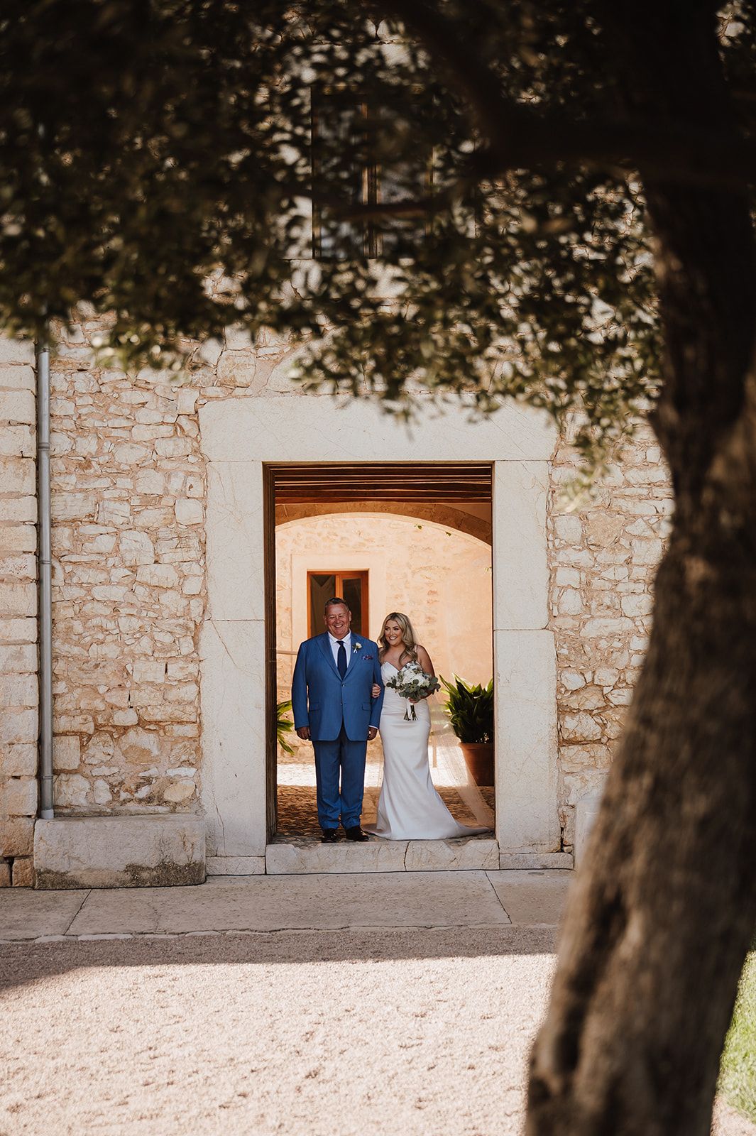 Bride and groom stand in stone archway doorway at elegant outdoor wedding venue surrounded by rustic walls and trees.