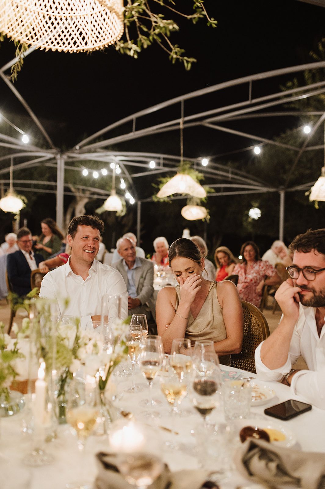 Wedding guests laughing and enjoying speeches at an elegant outdoor evening reception.