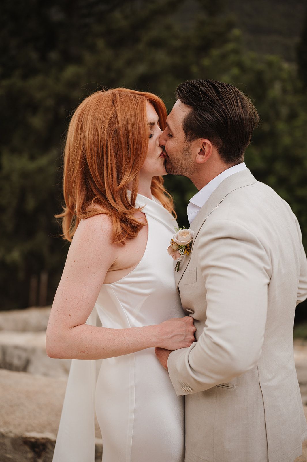 Bride with flowing red hair and groom in beige suit sharing a close romantic kiss outdoors.