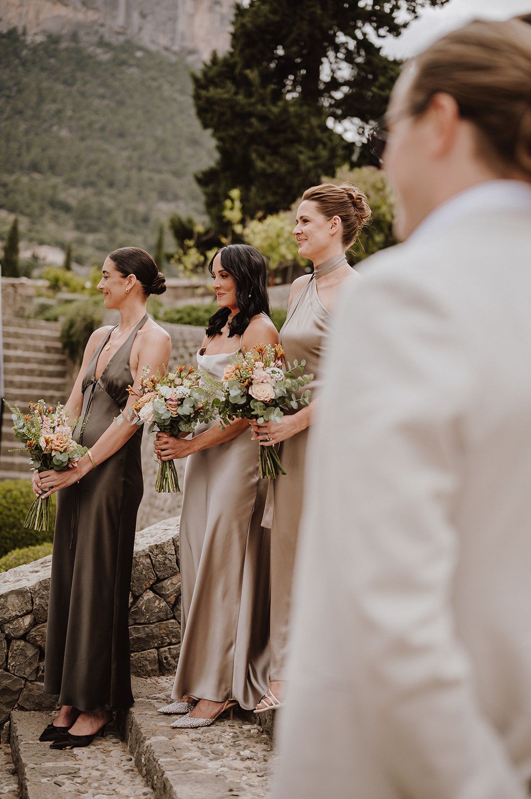 Three bridesmaids in satin halter gowns holding bouquets stand during an outdoor wedding ceremony.