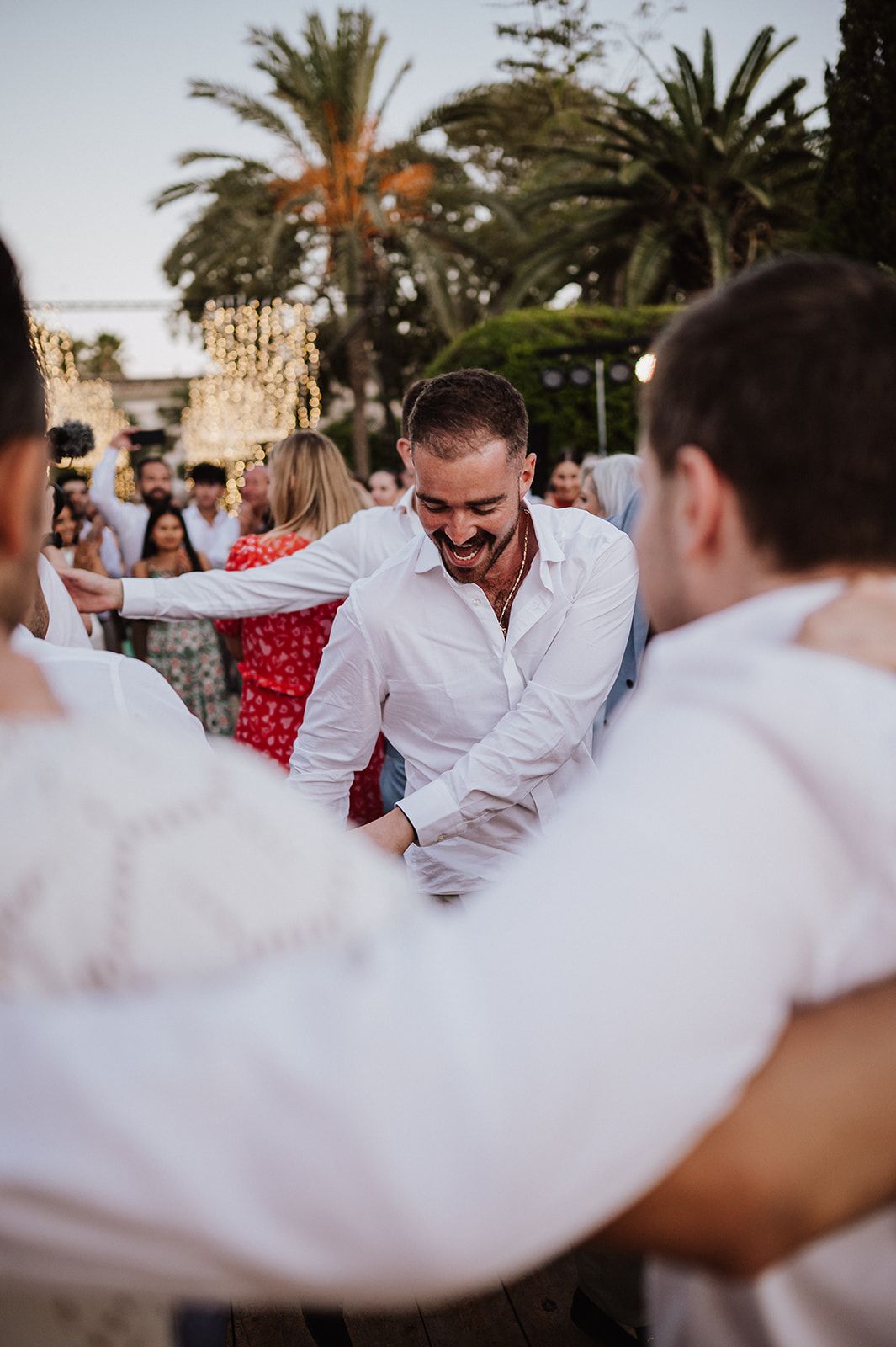 Groom in white shirt laughs joyfully while dancing with groomsmen in a circle at outdoor wedding reception.