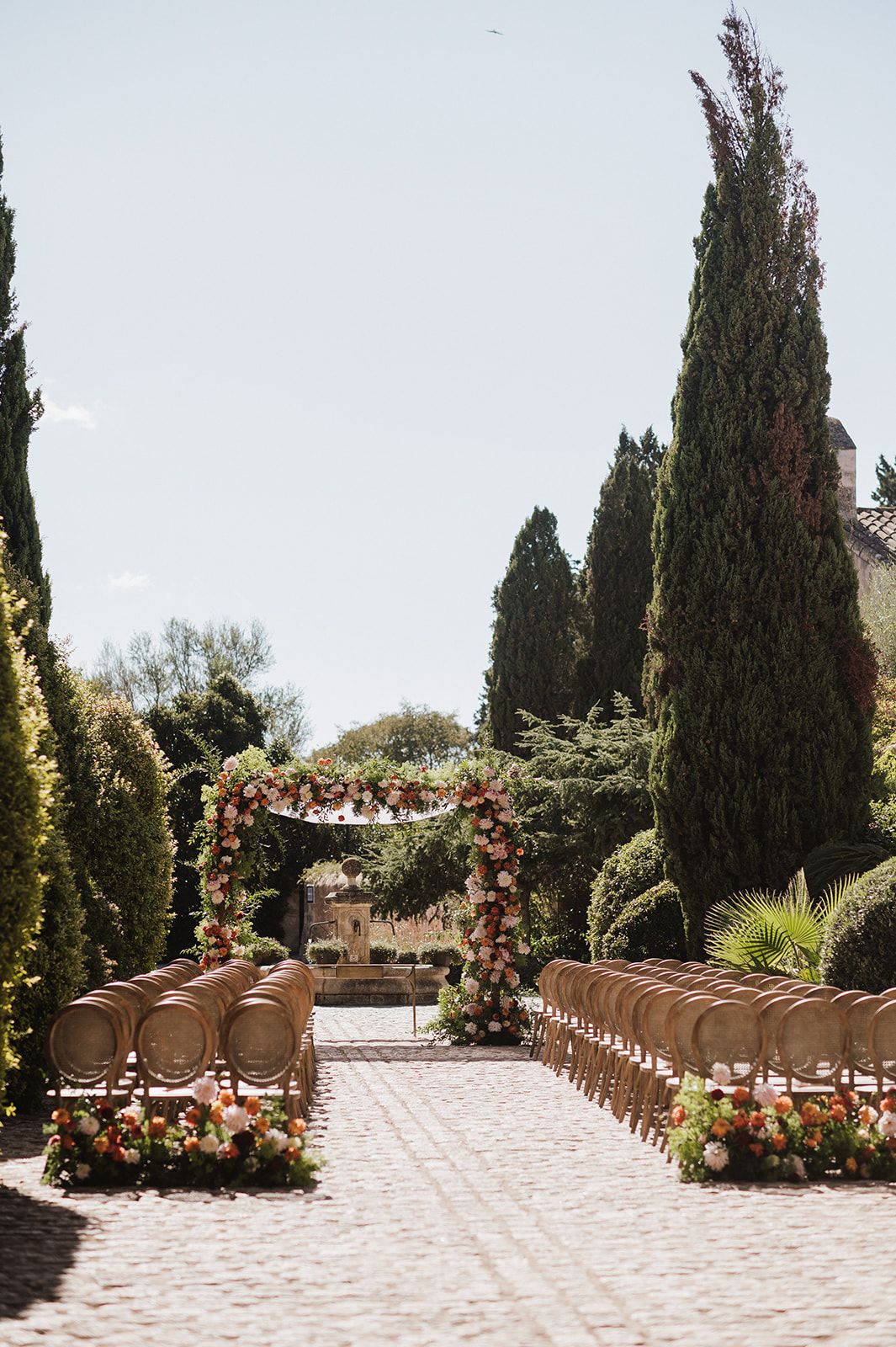 Elegant outdoor wedding ceremony aisle with gold chairs, lush floral arch, and tall cypress trees in warm sunlight.