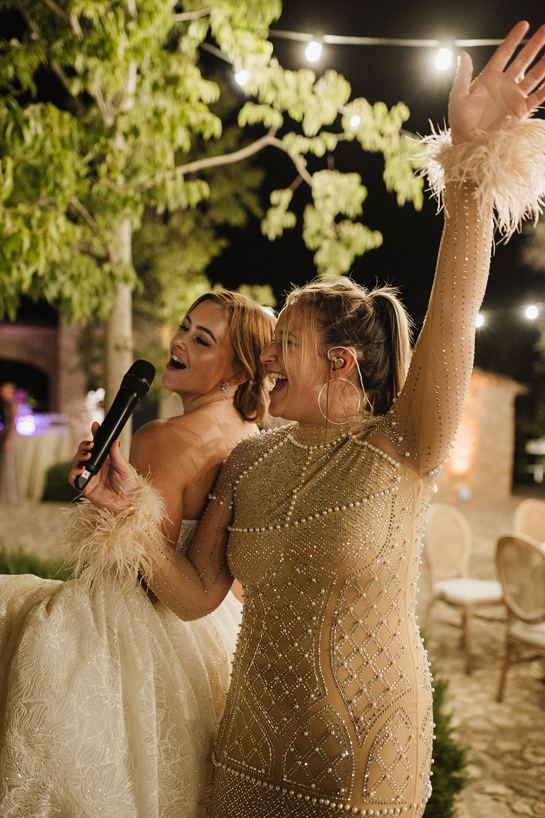 Two women in elegant dresses sing together at an outdoor wedding reception under string lights at night.