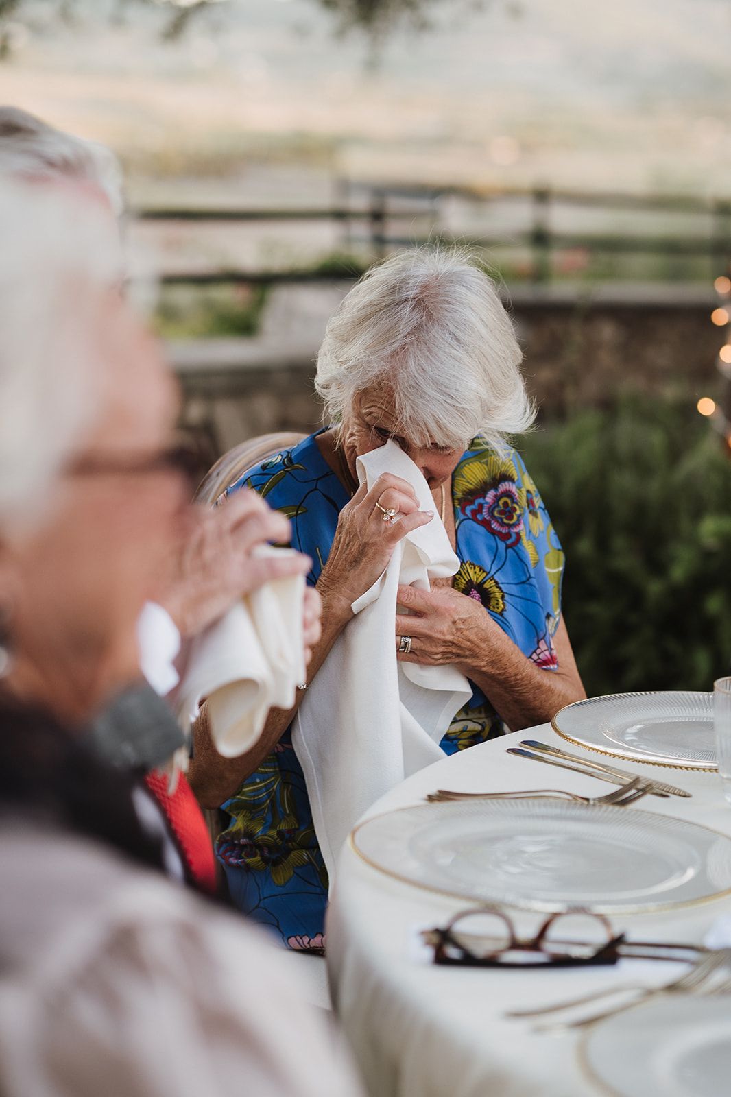 Two elderly women dab tears with napkins at an elegant outdoor wedding reception dinner table.