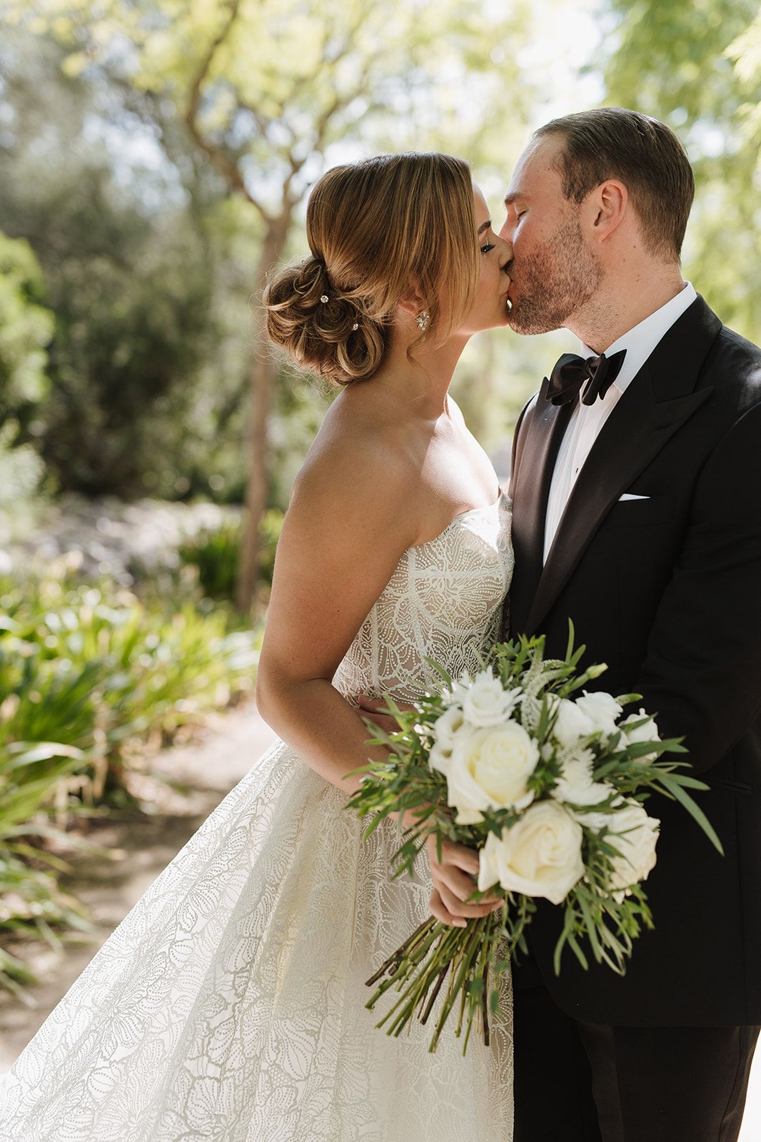Bride and groom share a kiss outdoors, bride holding white peony bouquet in lace gown, groom in black tuxedo.