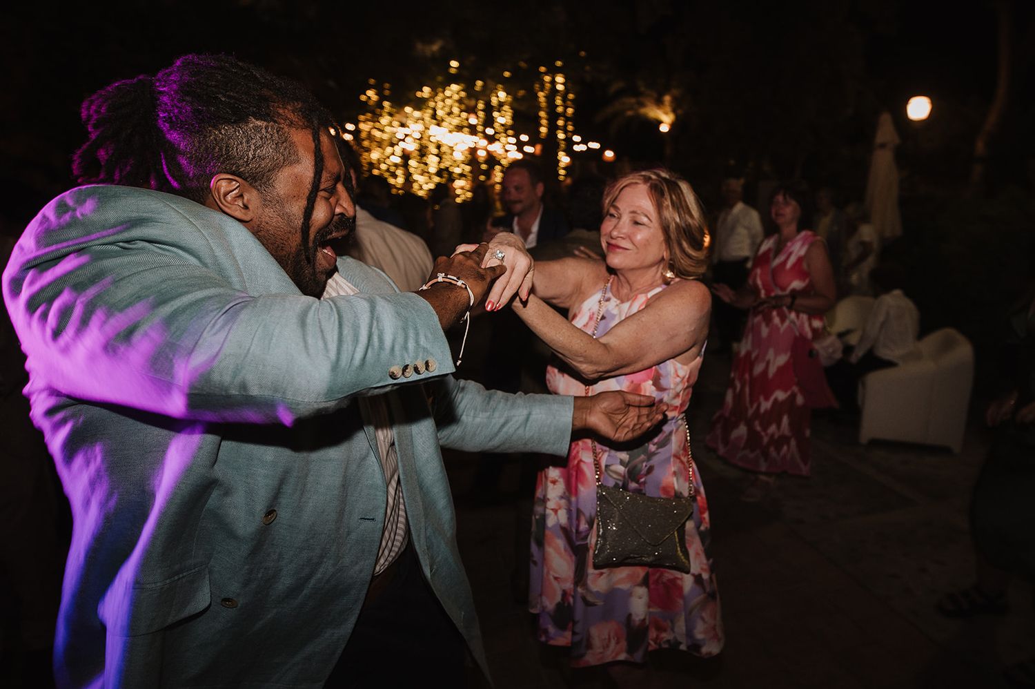Two wedding guests dancing energetically outdoors at night under warm string lights during wedding reception.