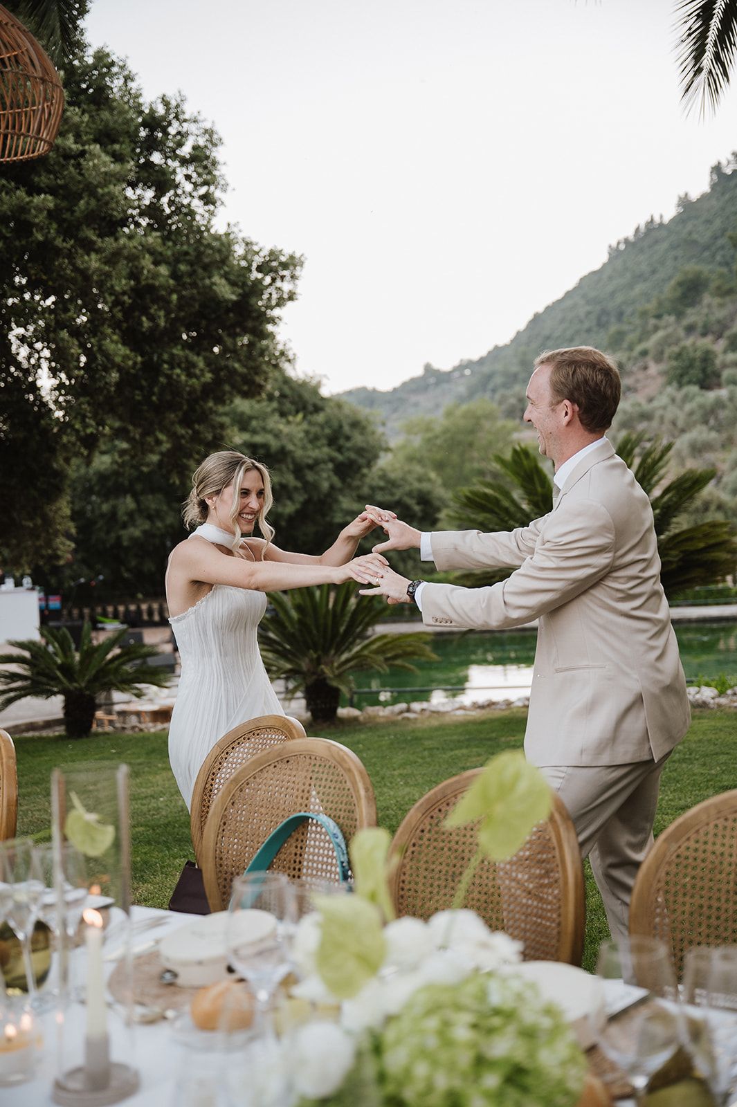 Bride and groom share a joyful dance outdoors with lush green mountains and palm trees in background.