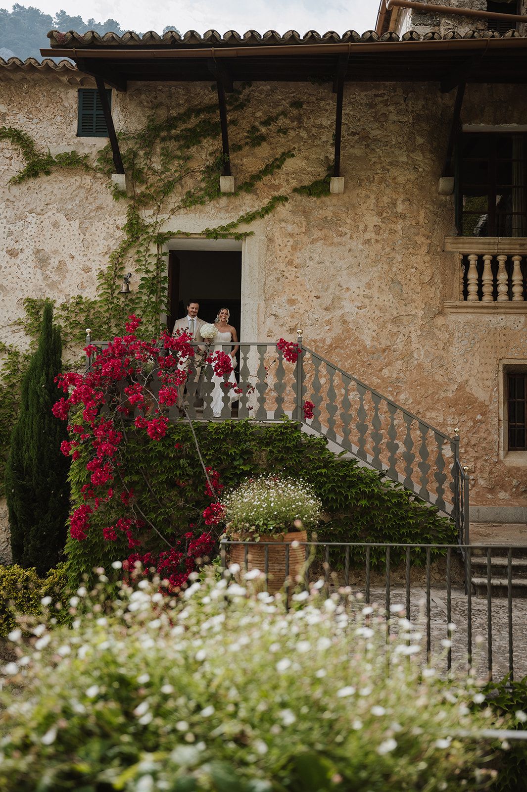 Couple stands on stone villa balcony surrounded by lush red bougainvillea and manicured gardens.