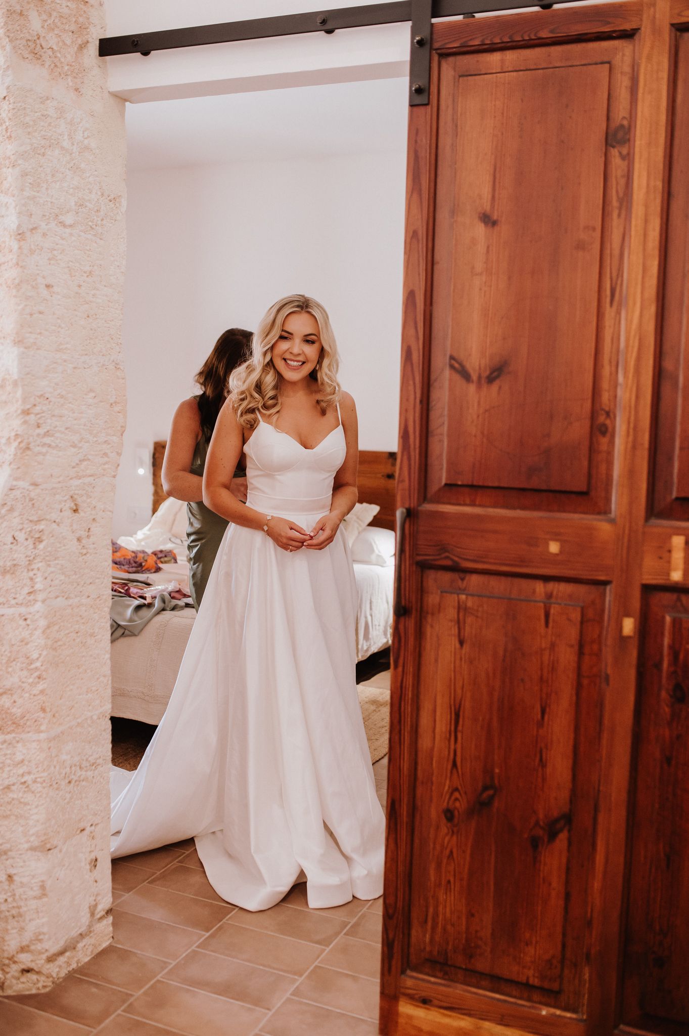 Smiling blonde bride in a simple white spaghetti strap wedding gown standing in a rustic doorway with barn door.