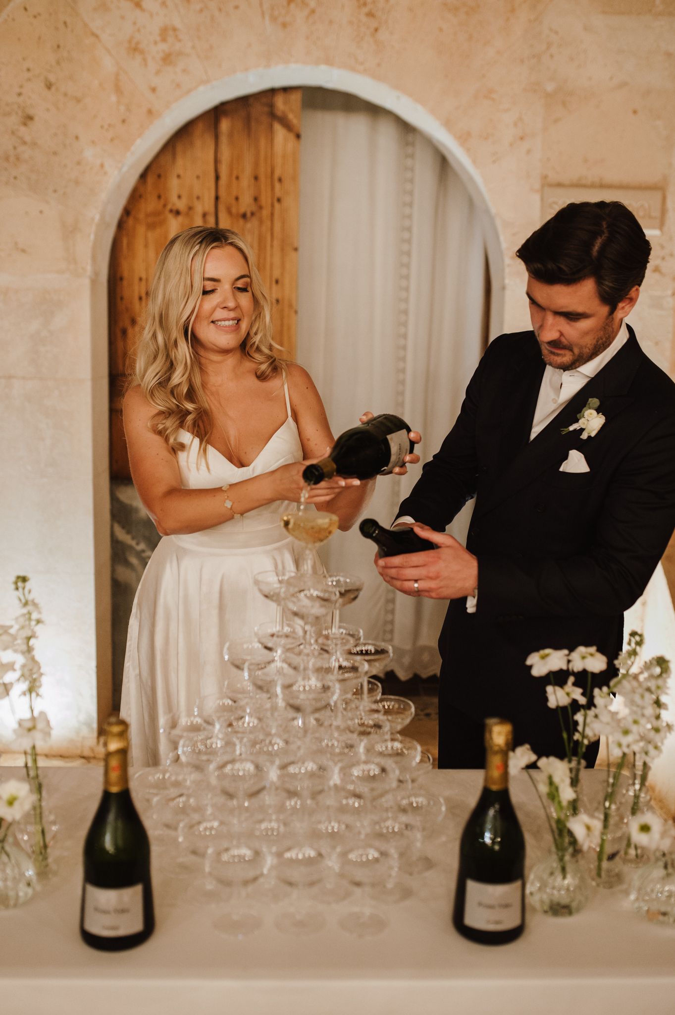 Smiling bride and groom pour champagne into a pyramid of glasses at their wedding reception.