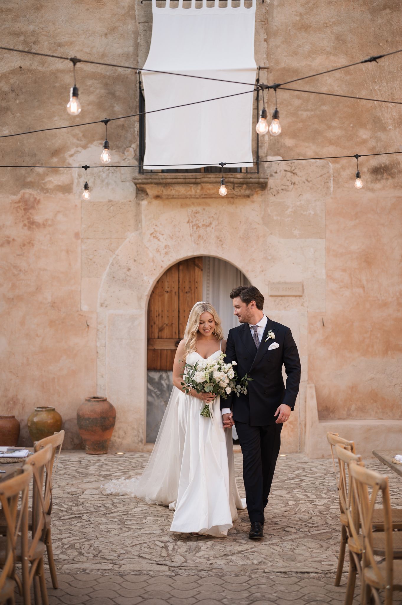 Newlyweds walking together through charming stone courtyard with string lights overhead.