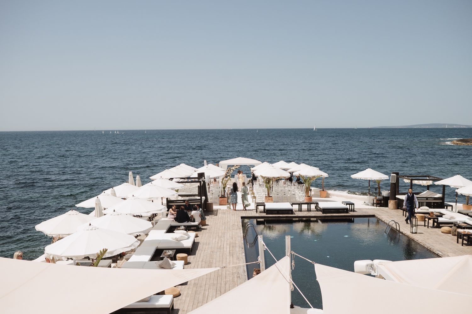 Aerial view of luxurious seaside wedding venue with white umbrellas and pool deck overlooking calm blue ocean.