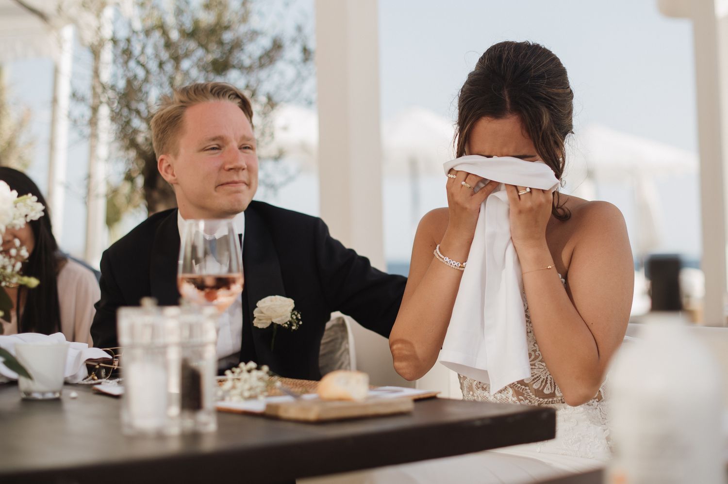 Emotional bride wiping tears with napkin while groom watches lovingly during outdoor seaside wedding reception.